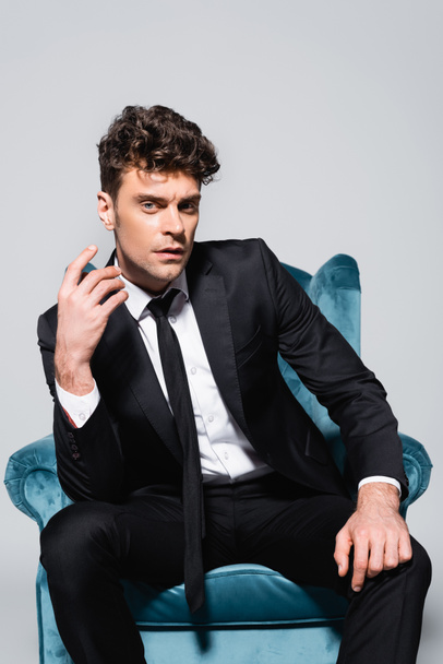 elegant man in suit sitting in velour armchair isolated on grey - 写真・画像