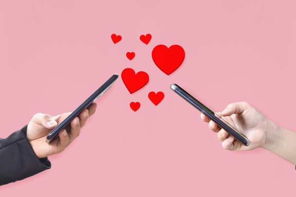 Mãos de casal segurando telefone celular com corações, símbolo de amor no fundo rosa. Conceito de dia dos namorados. - Foto, Imagem