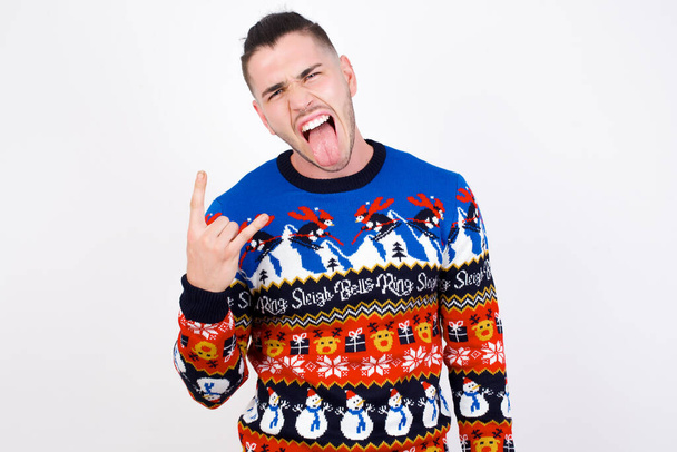 Портрет божевільного кавказького чоловіка в новорічному светрі на білій стіні, що показує язичкові роги вгору жестом, виражаючи хвилювання від перебування на концерті гурту. - Фото, зображення