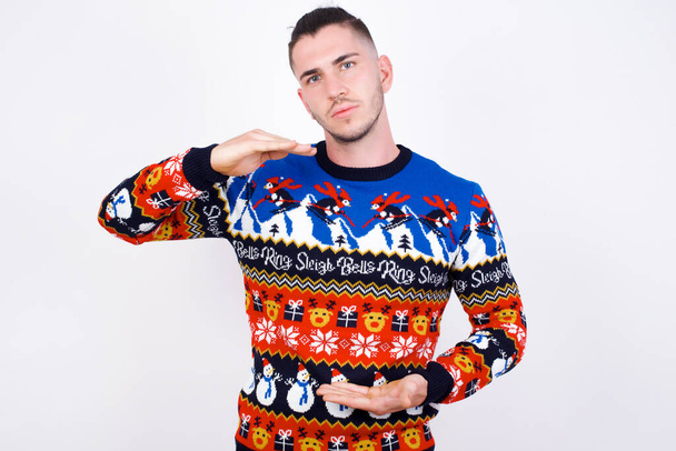 fehér férfi visel karácsonyi pulóver ellen fehér fal gesztikuláló kezek mutató nagy és nagy méretű jel, intézkedés szimbólum.  - Fotó, kép