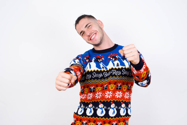 Портрет чарівного кавказького чоловіка в новорічному светрі на білій стіні, широко посміхаючись, тримаючи руки над головою. Впевненість і концепція розслаблення. - Фото, зображення
