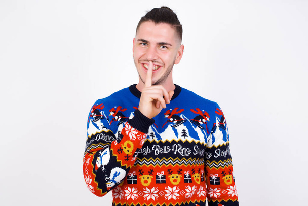 Χαμογελώντας νεαρός όμορφος Καυκάσιος άνδρας φορώντας χριστουγεννιάτικο πουλόβερ στον λευκό τοίχο, κάνει σουτ χειρονομία, κρατά το μπροστινό δάχτυλο πάνω από τα χείλη κρύβει μυστικό. Κάνε ησυχία, σε παρακαλώ.. - Φωτογραφία, εικόνα