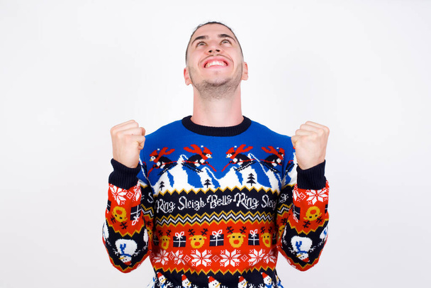 白い壁にクリスマスのセーターを着た若いハンサムな白人男性は、彼の目標を達成するために喜んで成功しています。勝利と勝利の概念. - 写真・画像