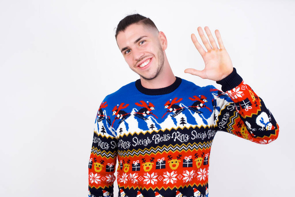 Kaukasische man draagt kersttrui tegen witte muur afzien van hallo zeggen of afscheid gelukkig en glimlachend, vriendelijk welkomstgebaar. - Foto, afbeelding