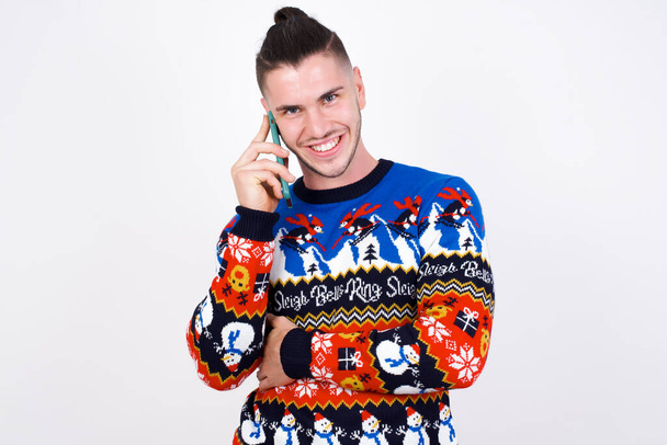 Portret van een lachende jonge knappe blanke man in een kersttrui tegen een witte muur, pratend op een mobiele telefoon. Bedrijfs-, vertrouwens- en communicatieconcept. - Foto, afbeelding