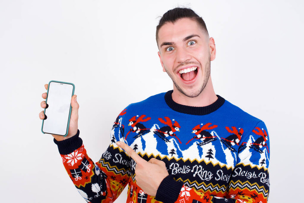 Excited Young handsome Caucasian man wearing Christmas sweater against white wall, holding and pointing with finger at smartphone with blank screen. Advertisement concept. - Photo, Image