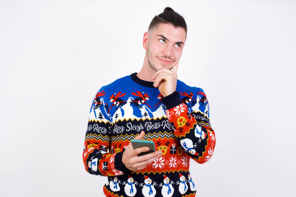 Pensando soñando joven guapo hombre caucásico usando suéter de Navidad contra la pared blanca, usando el teléfono móvil y sosteniendo la mano en la cara. Toma de decisiones y concepto de redes sociales. - Foto, imagen