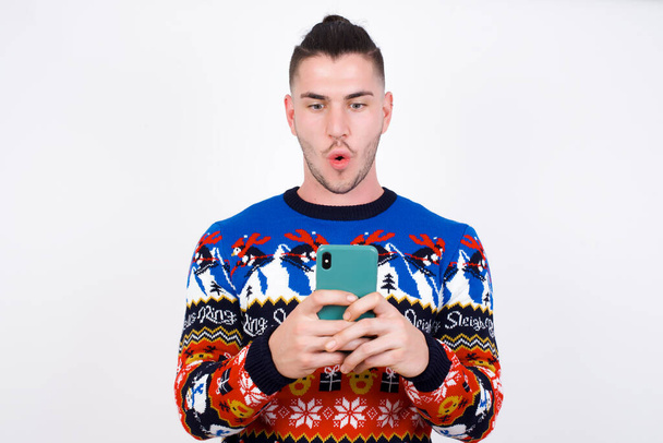 Joven hombre caucásico guapo que usa suéter de Navidad contra la pared blanca, mira con los ojos tapados, sostiene el teléfono inteligente moderno, recibe un mensaje inesperado de un amigo, lee el recordatorio. - Foto, imagen