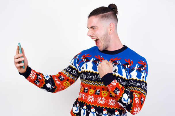 Счастливый молодой красивый белый мужчина в рождественском свитере на белой стене, получает хорошие новости по электронной почте и празднует успех, стоя и глядя на мобильный телефон. - Фото, изображение
