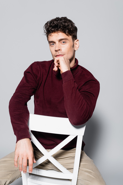 young man in burgundy jumper posing on chair isolated on grey - Foto, Imagem