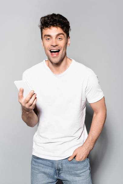 happy young man in white t-shirt with smartphone isolated on grey - 写真・画像