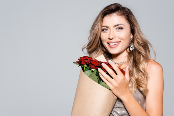 mujer elegante sonriente en vestido con ramo de rosas rojas aisladas en gris - Foto, imagen