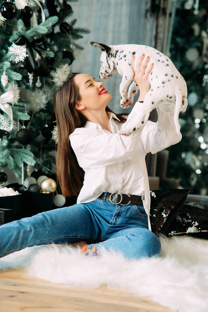Νεαρή γυναίκα κάθεται στο πάτωμα κοντά στο χριστουγεννιάτικο δέντρο με διακοσμήσεις και παίζει με το κουτάβι του Δαλματικού σκύλου. - Φωτογραφία, εικόνα