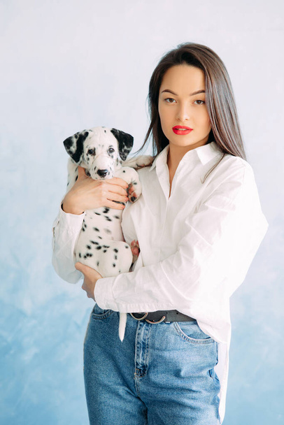 Junge Frau steht mit Welpe eines Dalmatinerhundes in der Hand vor blauem Hintergrund. - Foto, Bild