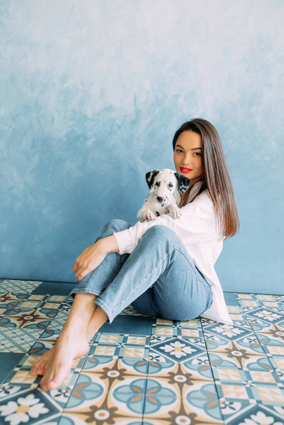 Young woman sits with puppy of dalmatian dog in her hands against blue background. - Фото, зображення