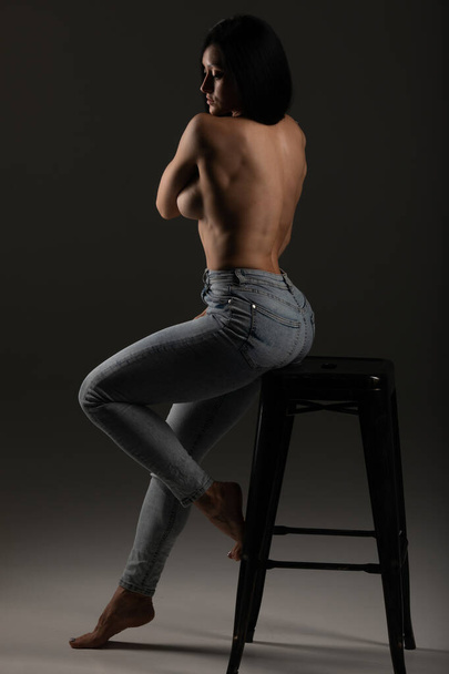 Sexy fit brunette in jeans sitting on chair showing strong back muscles. - Photo, Image
