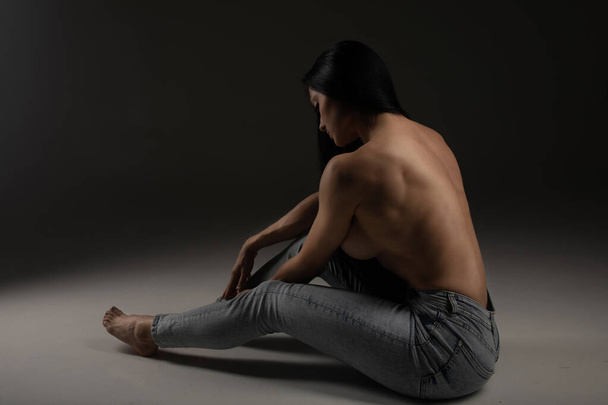 Сексуальна без сорочки брюнетка жінка в джинсах позує сидячи на підлозі студії
 - Фото, зображення
