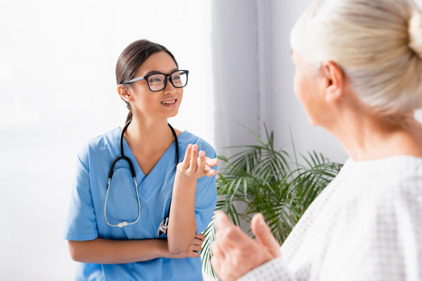 sonriente asiático enfermera en gafas gesto mientras hablando a senior mujer en hospital - Foto, imagen