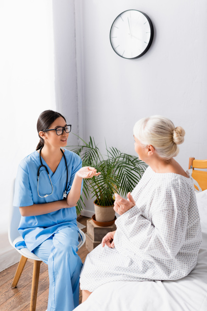 giovane infermiera asiatica gesticolando mentre seduto e parlando con la donna anziana in ospedale - Foto, immagini