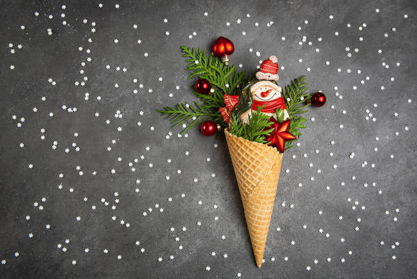 Snowman, thuja twigs and Christmas toys in a waffle cone. Dark gray background with snowflakes. Christmas card. An original sweet gift. - Zdjęcie, obraz