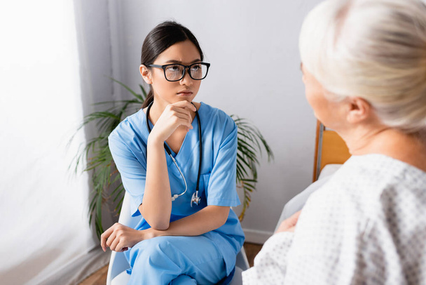ernstige aziatische verpleegkundige kijken naar oudere vrouw tijdens gesprek in het ziekenhuis, wazig voorgrond - Foto, afbeelding