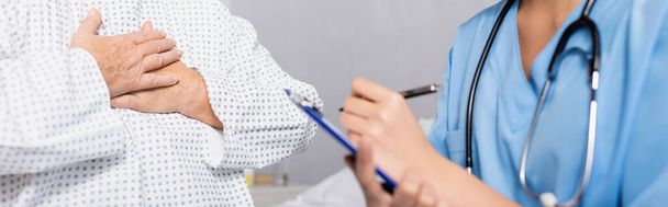 cropped view of senior woman touching chest while suffering from gasp attack near nurse writing on clipboard, blurred foreground, banner - Photo, Image