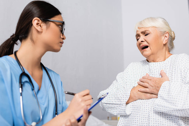 věk žena dotýkající se hrudníku, zatímco trpí astmatickým záchvatem v blízkosti asijské zdravotní sestry psaní na schránce, rozmazané popředí - Fotografie, Obrázek