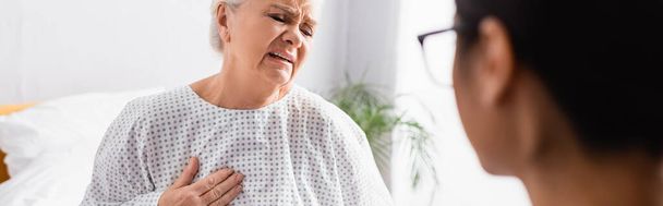 mujer mayor tocando el pecho mientras sufre de dolor de corazón cerca de la enfermera en primer plano borrosa, bandera - Foto, Imagen