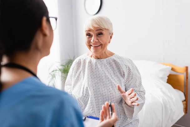 happy senior woman gesturing while talking to nurse in hospital, blurred foreground - Фото, изображение