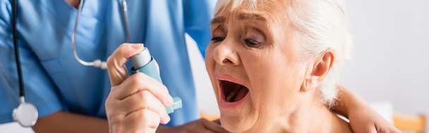 mujer de edad avanzada con la boca abierta que sufre de ataque de asma y el uso de inhalador cerca de la enfermera sobre fondo borroso, bandera - Foto, Imagen