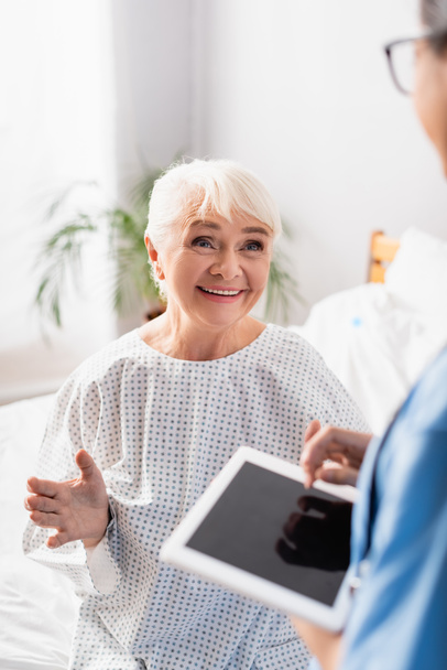 happy aged woman gesturing while talking to nurse holding digital tablet with blank screen on blurred foreground - Fotó, kép