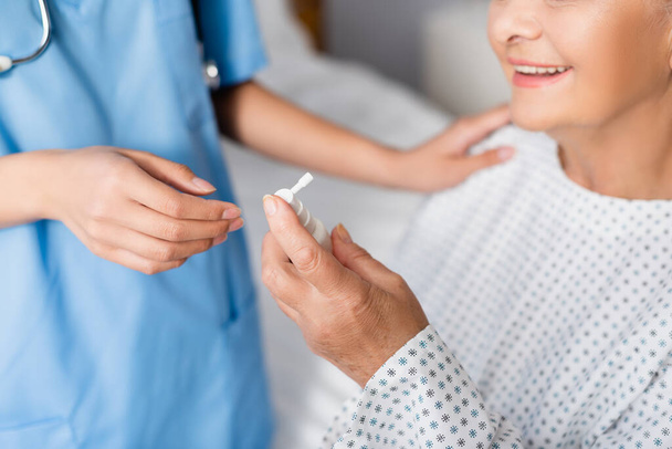 cropped view of aged, smiling woman holding nasal spray near nurse on blurred background - Photo, Image