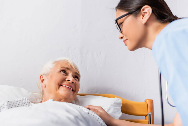young asian nurse looking at smiling elderly woman lying in hospital - Фото, зображення