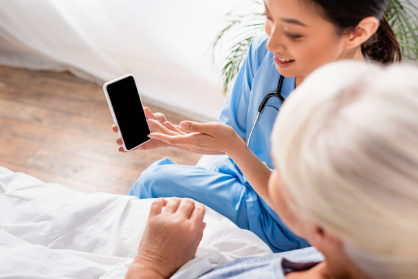 overhead view of smiling asian nurse pointing at mobile phone near senior woman on blurred foreground - Foto, imagen