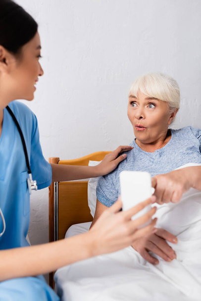 amazed elderly woman pointing at smartphone near young asian nurse, blurred foreground - Фото, изображение