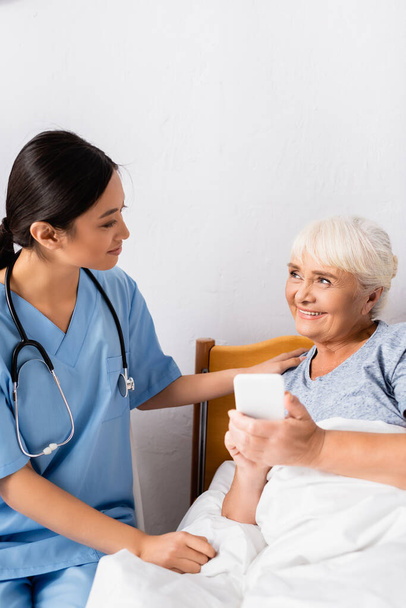χαρούμενη ηλικιωμένη γυναίκα που κοιτάζει χαμογελαστή ασιάτισσα νοσοκόμα ενώ κρατά το smartphone με κενή οθόνη - Φωτογραφία, εικόνα