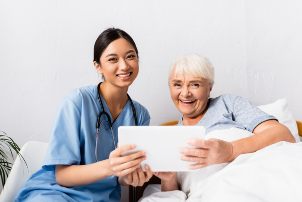 glückliche asiatische Krankenschwester und lachende Seniorin schaut in die Kamera, während sie ein digitales Tablet in der Hand hält - Foto, Bild