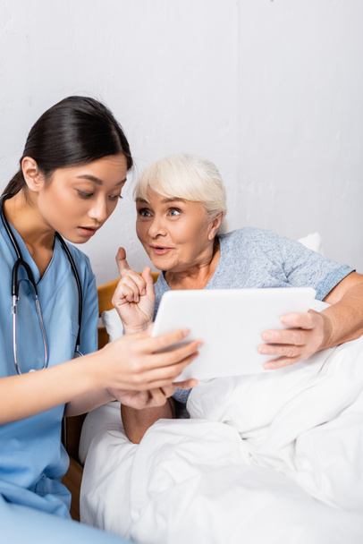 aufgeregte Seniorin zeigt Ideengeste beim Umgang mit digitalem Tablet zusammen mit staunender asiatischer Krankenschwester  - Foto, Bild