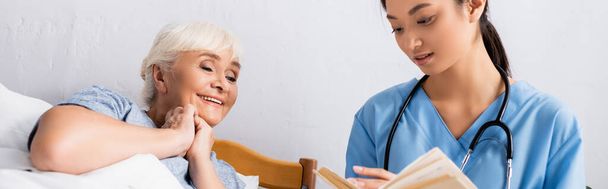 young asian nurse reading book to happy aged woman in hospital, banner - Photo, Image