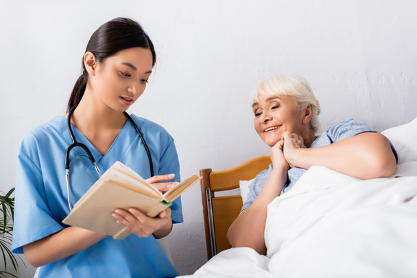 asiático enfermera lectura libro a alegre anciano mujer en hospital - Foto, imagen