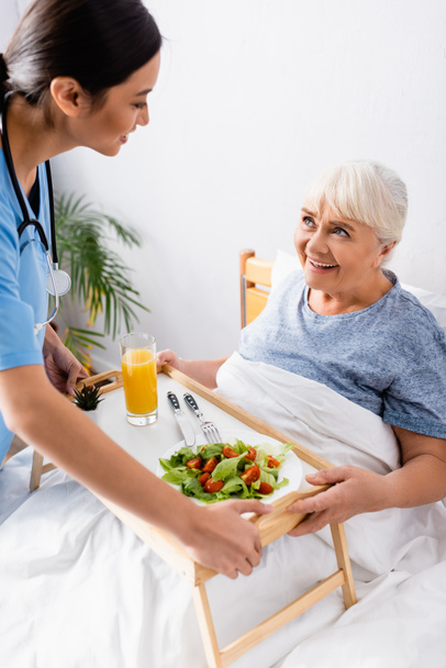 smiling asian nurse holding tray with breakfast near happy elderly woman in bed - Foto, afbeelding