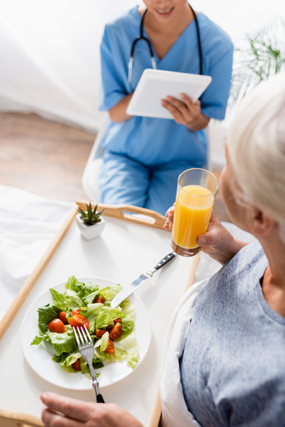 vysoký úhel pohledu starší ženy se snídaní v blízkosti zdravotní sestry pomocí digitální tablet na rozmazaném pozadí - Fotografie, Obrázek