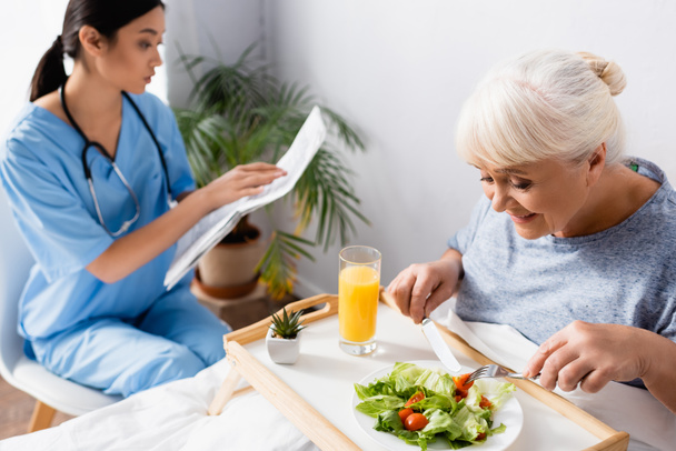 pleased senior woman having breakfast near young asian nurse reading newspaper on blurred background - Photo, Image
