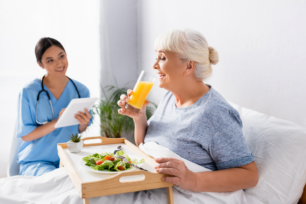 pleased aged woman having breakfast and drinking orange juice near smiling asian nurse on blurred background - Фото, изображение