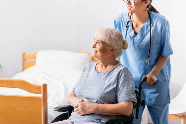 young nurse near smiling aged woman sitting in wheelchair and looking away - Photo, Image