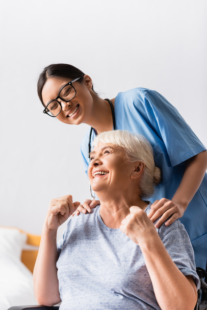nízký úhel pohledu na vzrušený starý žena ukazující vítězný gesto v blízkosti šťastný asijské zdravotní sestra - Fotografie, Obrázek