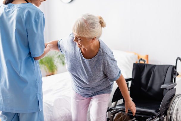 junge Krankenschwester hilft alter Frau beim Aufstehen aus dem Rollstuhl - Foto, Bild