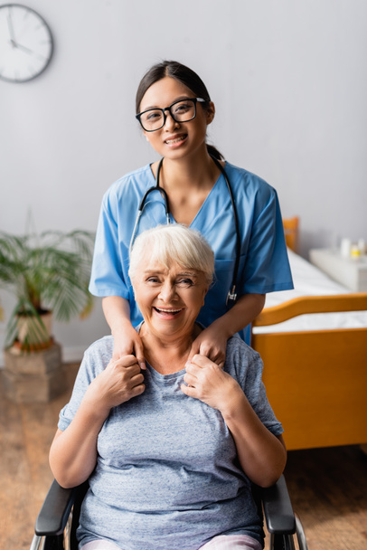 счастливая азиатская медсестра и смеющаяся пожилая женщина в инвалидном кресле держась за руки, глядя в камеру - Фото, изображение