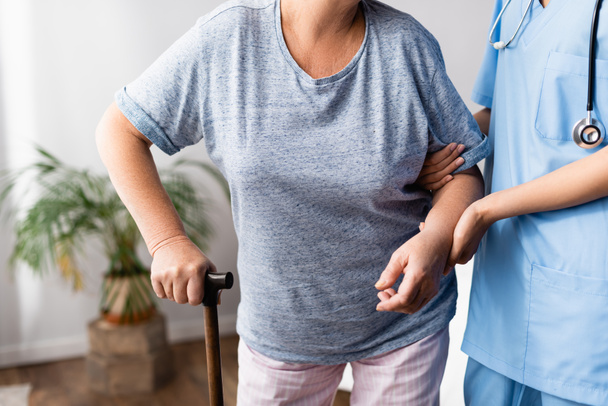 gedeeltelijk zicht op verpleegkundige ondersteuning van oudere vrouw met wandelstok in het ziekenhuis - Foto, afbeelding