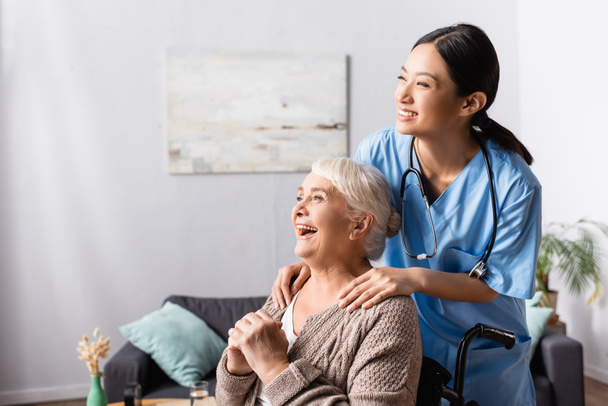 щаслива азіатська медсестра торкається плечей літньої інвалідної жінки, яка сміється в інвалідному візку
 - Фото, зображення